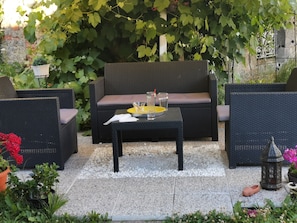 An outdoor seating area with wicker furniture, a small table with a glass and a yellow tray, surrounded by greenery and a decorative lantern. - Presbytere (Sauville)