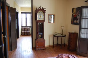 Una habitación con piso de madera, un reloj de pie, una mesita con una lámpara, una silla y una puerta.