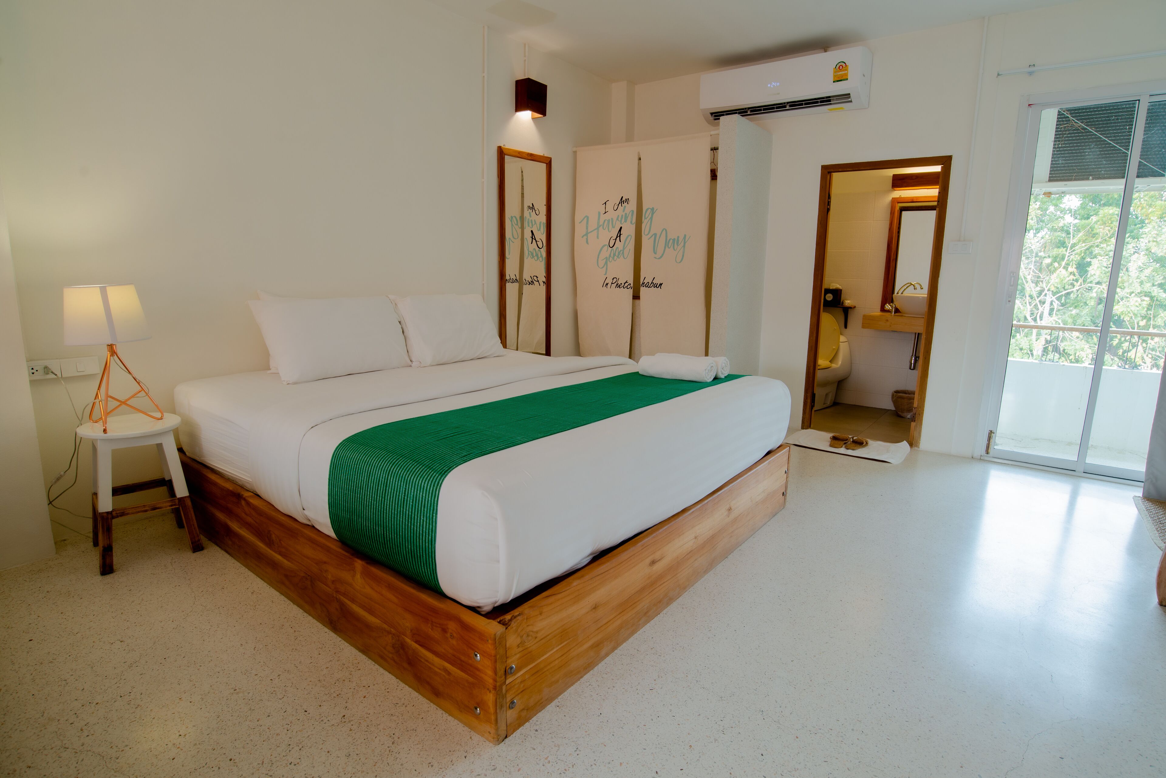 A bedroom with a wooden bed frame, white bedding, a green throw, a bedside lamp, and a view of the outdoors through a sliding door.