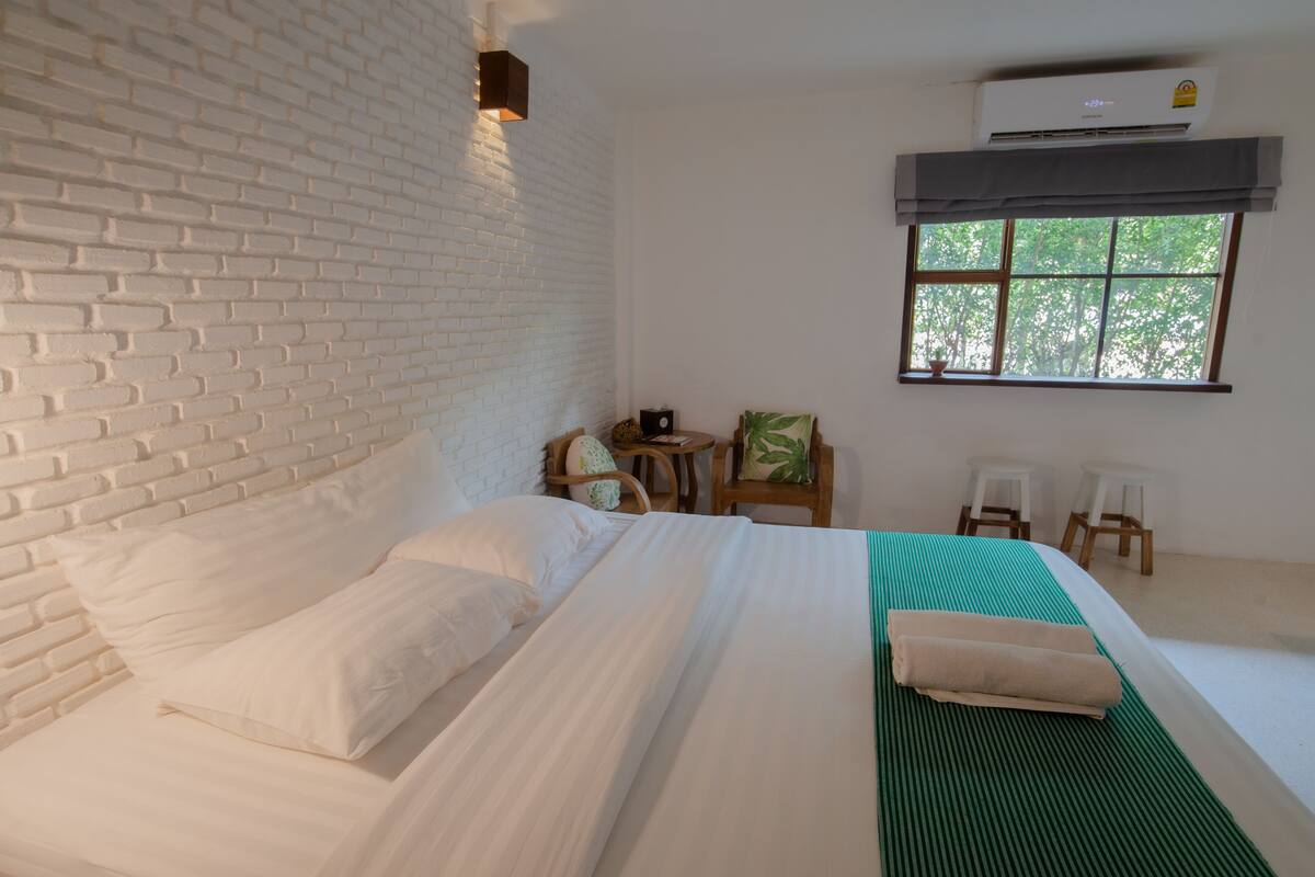 A room with a brick-patterned headboard, a bed with white linens, a green mat, a small table with a chair, and a window with a view of trees.