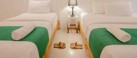 A hotel room with two beds, each with a green-striped bedspread and white pillows. There is a small table with a lamp between the beds, and a pair of slippers on the floor.