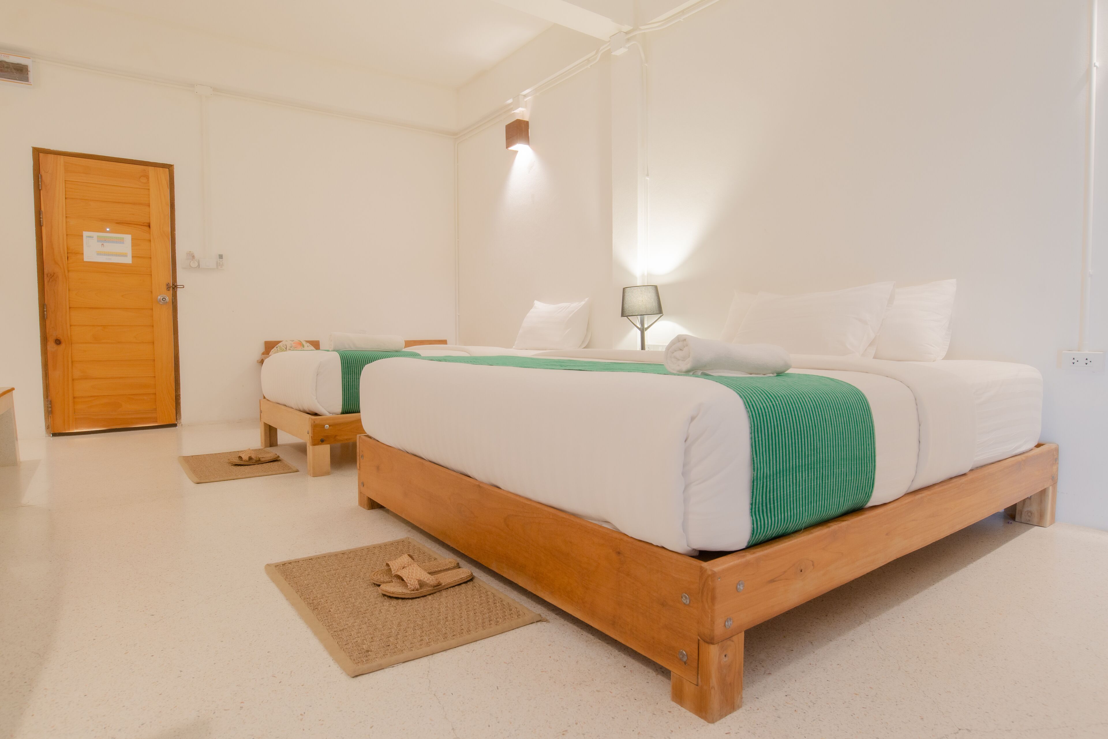 Une pièce avec deux lits en bois, une porte en bois, un petit tapis et une table de chevet avec une lampe.