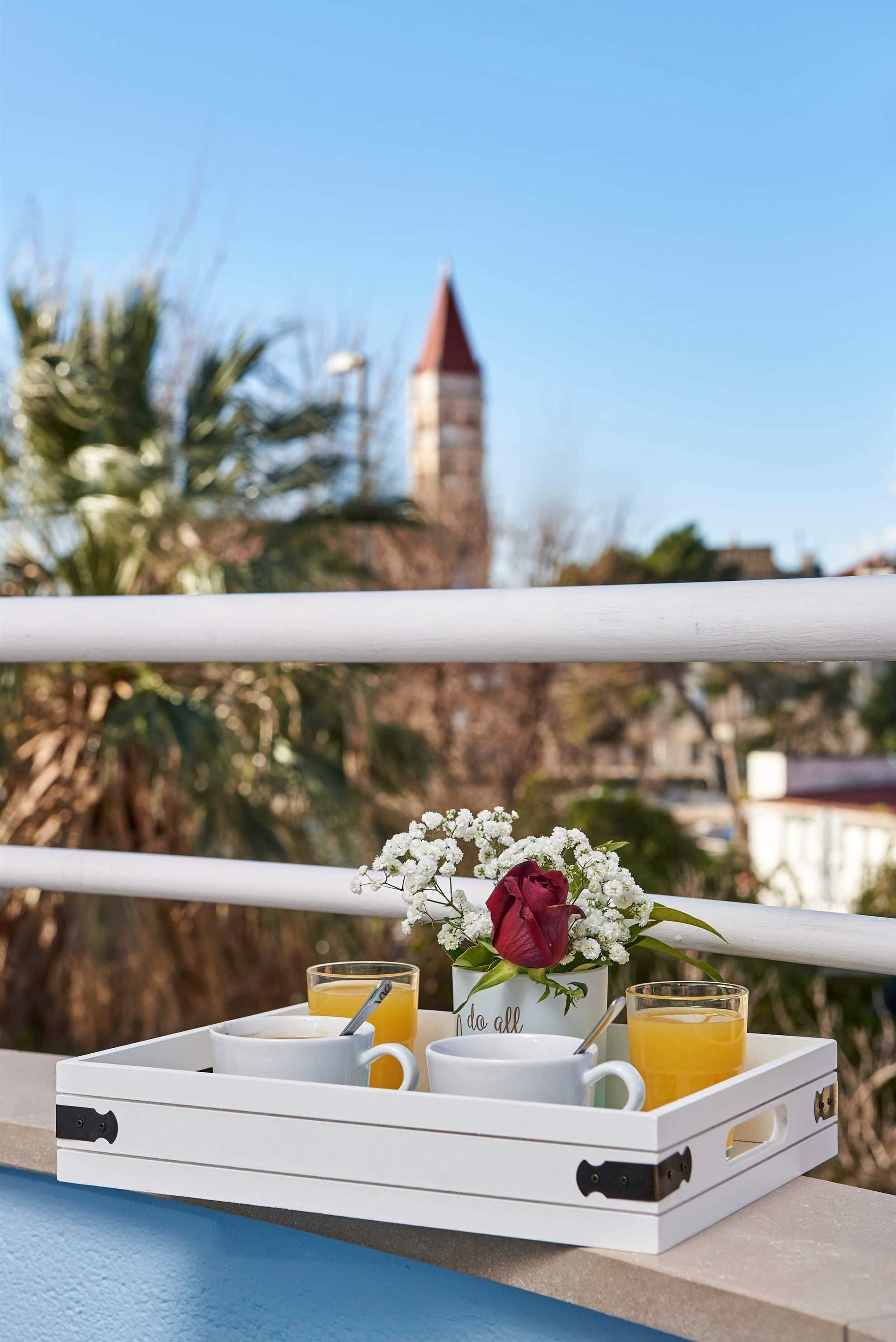 A tray with a flower arrangement, a red rose, and two glasses of orange juice on a balcony with a clear view of a building and palm trees.