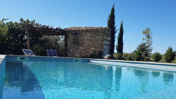 Una piscina trasparente con una struttura in pietra e un pergolato sullo sfondo.