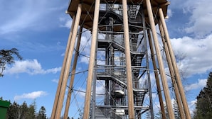 A tall, multi-level tower with wooden beams and metal railings, surrounded by a fenced area and trees. - Untere Kapfenhardter Mühle (Schoemberg)