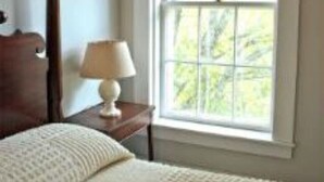 Quarto com uma janela ampla, cabeceira de madeira, mesa de cabeceira com abajur e cama com cobertor branco.
