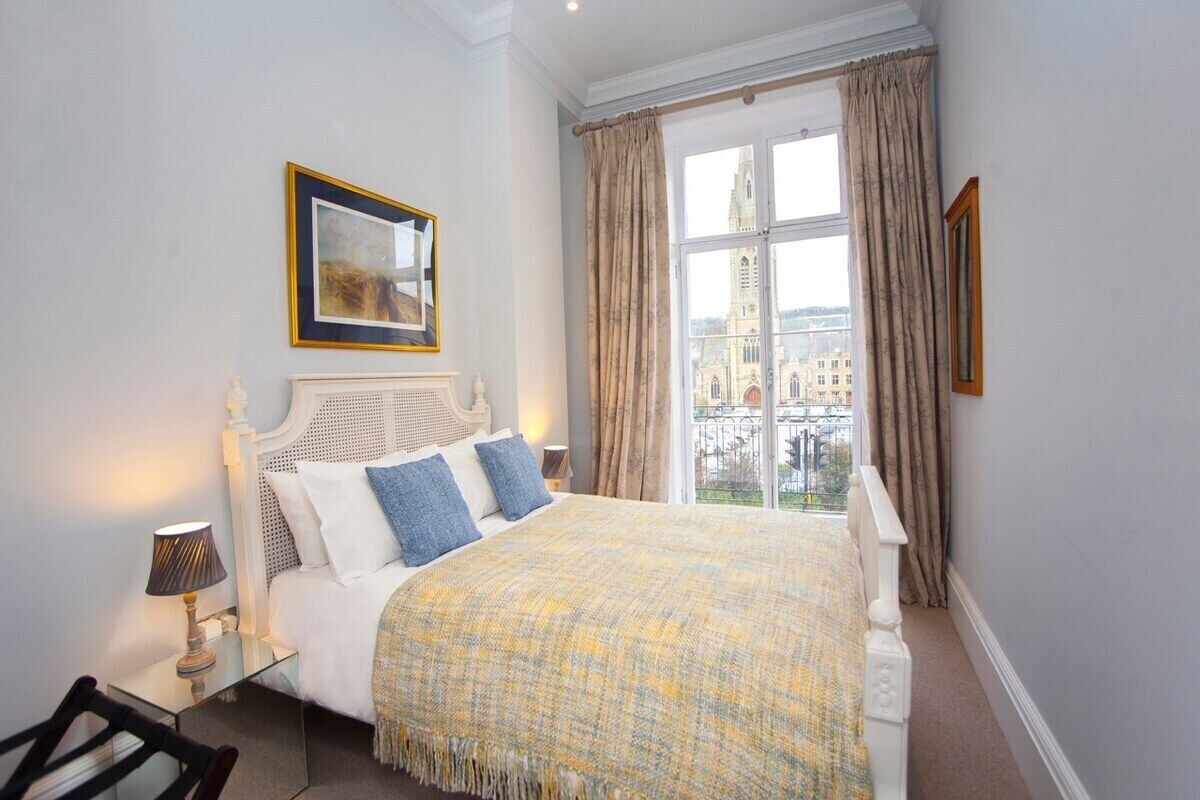 A bedroom with a large bed, a bedside table with a lamp, a framed picture on the wall, and a window with curtains.