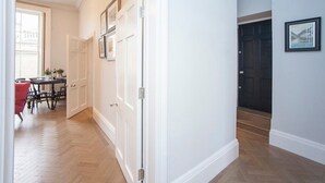 Un couloir avec un plancher en bois, des murs blancs et une porte fermée. Un tableau encadré est accroché au mur, et une chaise rouge se trouve au fond.
