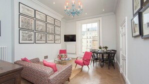 Un salon avec un canapé à motifs, une petite table avec une lampe, un coin repas avec une table et des chaises, et un lustre.