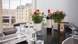 Une table à manger est dressée avec des assiettes, des couverts, des verres et une bouteille de vin Baracca Chianti. Deux plantes en pot, ornées de fleurs rouges, sont posées sur la table.