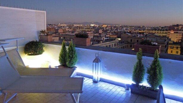A rooftop terrace with a lounge chair, potted plants, and a view of a cityscape at dusk. - Luxury 10thfloor Penthouse Stunning View Over Rome. Big Terrace (Roma)