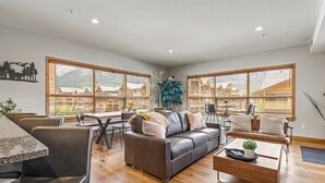 A spacious living room with a gray sofa, a wooden coffee table, and a dining area with a table and chairs. Large windows offer a view of a mountainous landscape. - Spring Creek 3F 1600sf Corner 3BR/2BTH (Canmore)