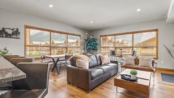 Amplia sala de estar con un sofá gris, una mesa de centro de madera y un comedor con mesa y sillas. Amplias ventanas ofrecen vistas a un paisaje montañoso.