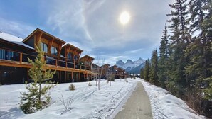 A snow-covered pathway leading to a building with wooden architecture, surrounded by trees and mountains in the background. - Spring Creek 3F 1600sf Corner 3BR/2BTH (Canmore)
