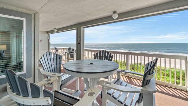 A covered outdoor seating area with Adirondack chairs and a table, overlooking the ocean.