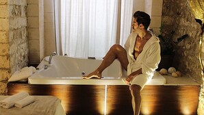 Un homme est assis dans une baignoire, dans une pièce où se trouvent un banc en bois, un lavabo et un pot de fleurs.