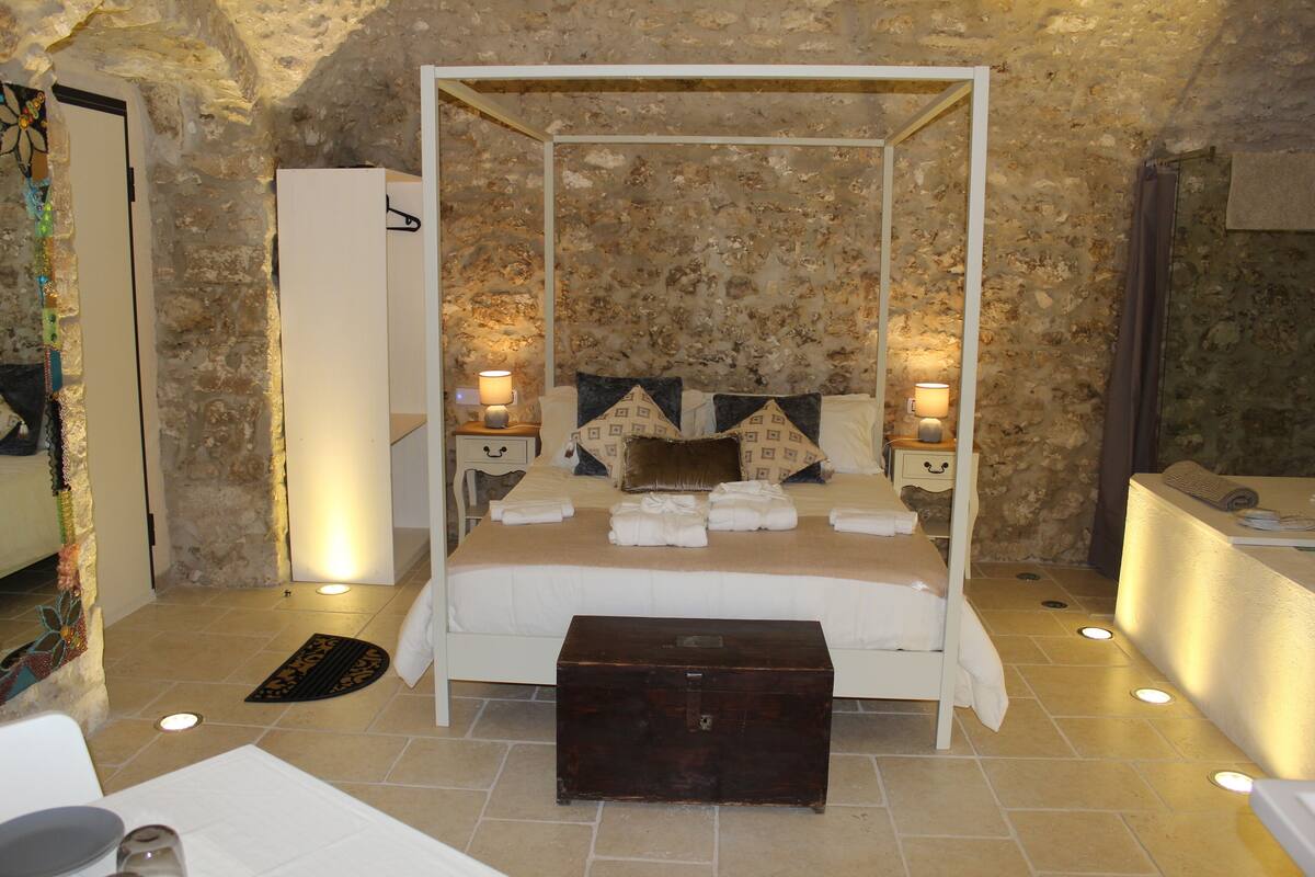 Un lit à baldaquin avec un mur en pierre en arrière-plan, des tables de chevet avec des lampes et une commode en bois dans une pièce dont le sol est en pierre.