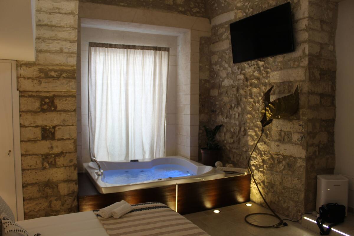 Une pièce avec un mur en pierre, une grande fenêtre avec des rideaux, une baignoire, un lit et un téléviseur à écran plat fixé au mur.