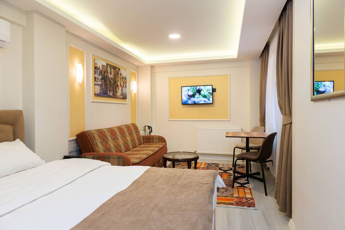 Una habitación de hotel con cama, sofá, una mesita, televisión y ventana con cortinas.
