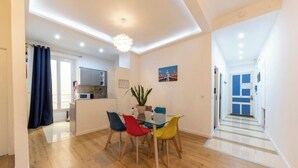 Un comedor moderno con una mesa de vidrio y sillas coloridas, una planta en maceta, y vistas de la cocina y el pasillo.
