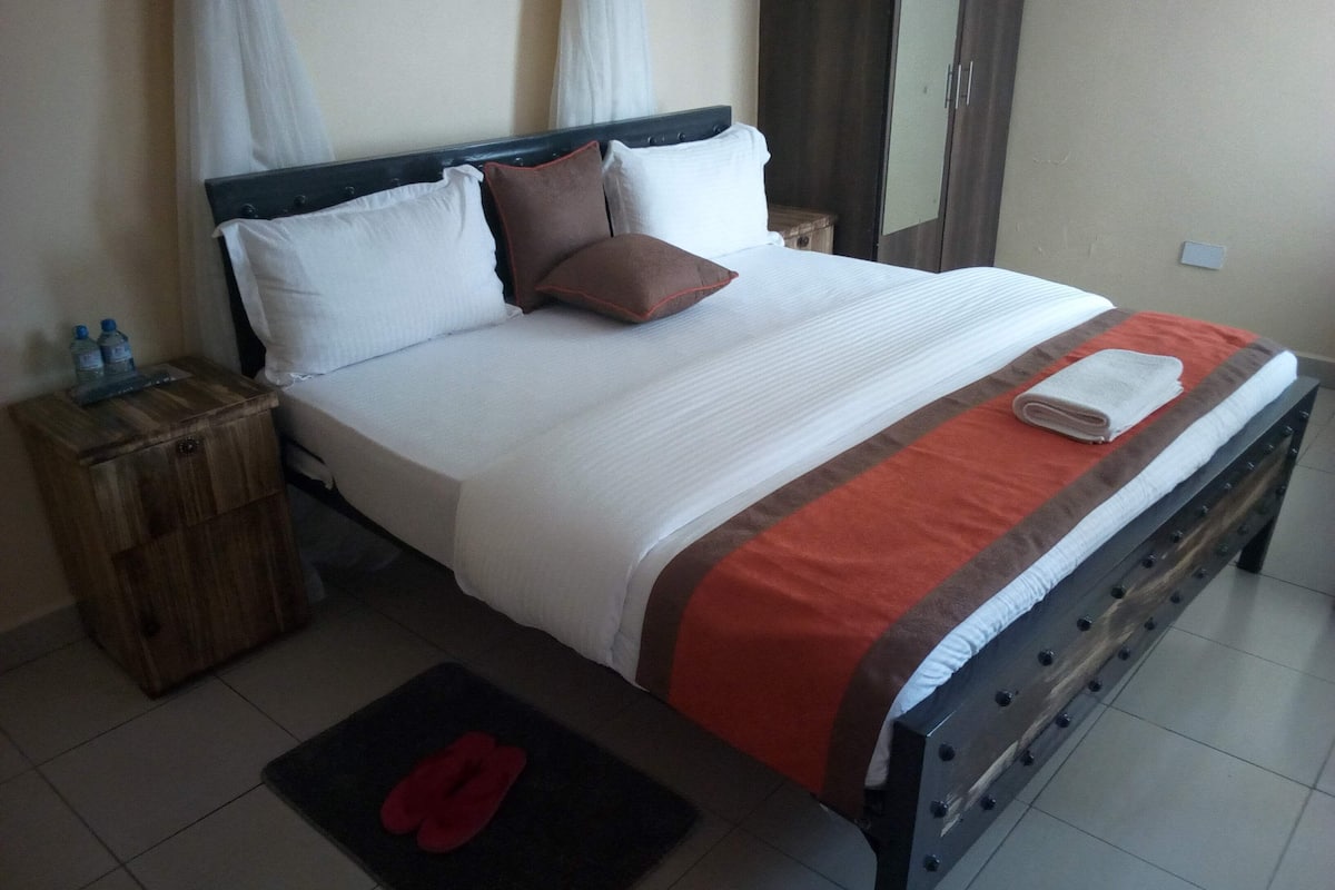 A neatly made bed with a white and brown bedspread, a wooden nightstand with a bottle of water, and a red rug on the floor.