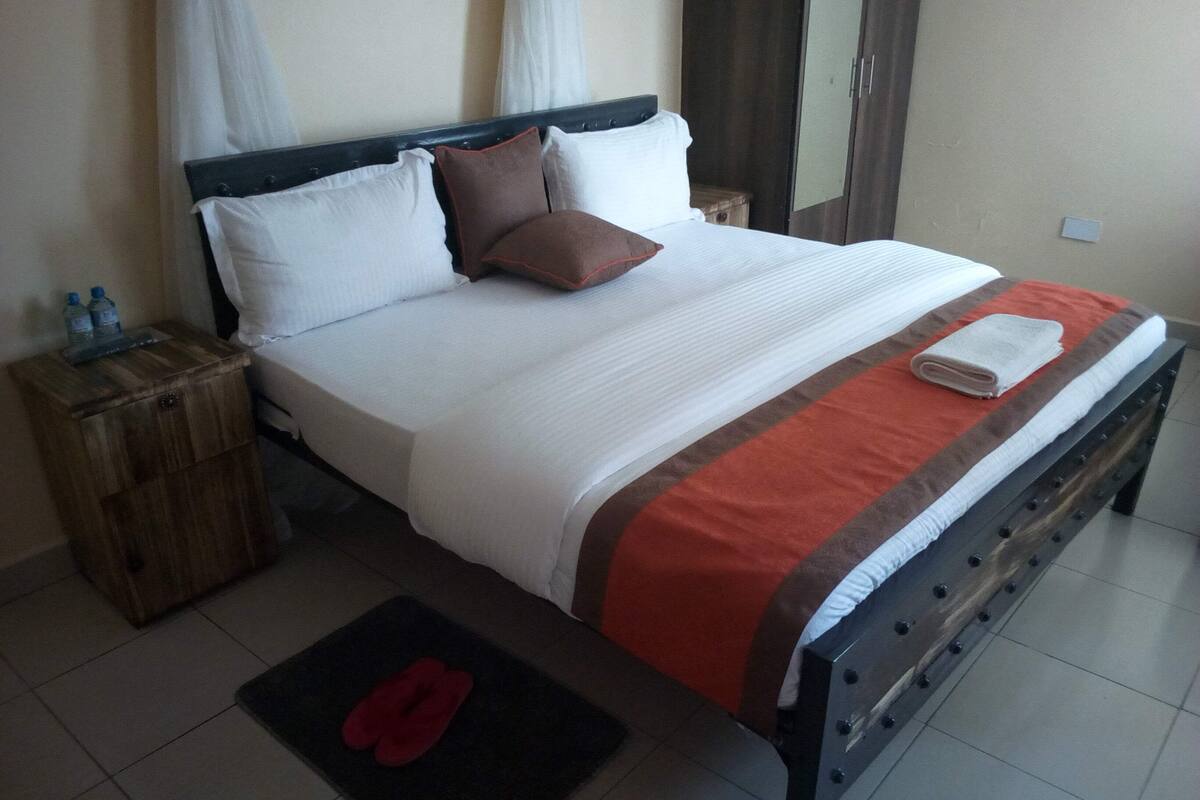 A neatly made bed with pillows, a wooden nightstand with bottles, and a red mat on the floor.