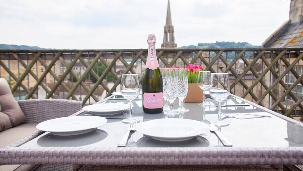 Mesa de comedor dispuesta con platos, vasos y una botella de champán Lanson, con vista a un paisaje urbano que incluye una aguja de iglesia.