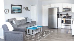 A modern living room with a grey sofa, a matching armchair, a coffee table, and a kitchen area with a fridge, oven, and microwave. - Gulf Views Across The Street From Beach Access (Madeira Beach)