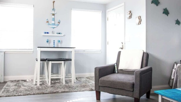 A room with a grey armchair, a white table with chairs, a decorative anchor, and a window with blinds. - Gulf Views Across The Street From Beach Access (Madeira Beach)