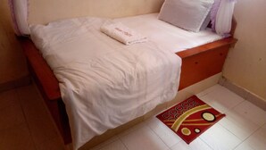 A single bed with a white mattress and a matching pillow, a small bedside table, and a red and yellow patterned rug on a tiled floor. - Ruby G Hotel and Club (Ruiru)