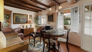 A dining area with a round glass table, wooden chairs, and a chandelier. There are two windows with white frames and a view of greenery outside. - Quinta Florença - By Wehost (Funchal)