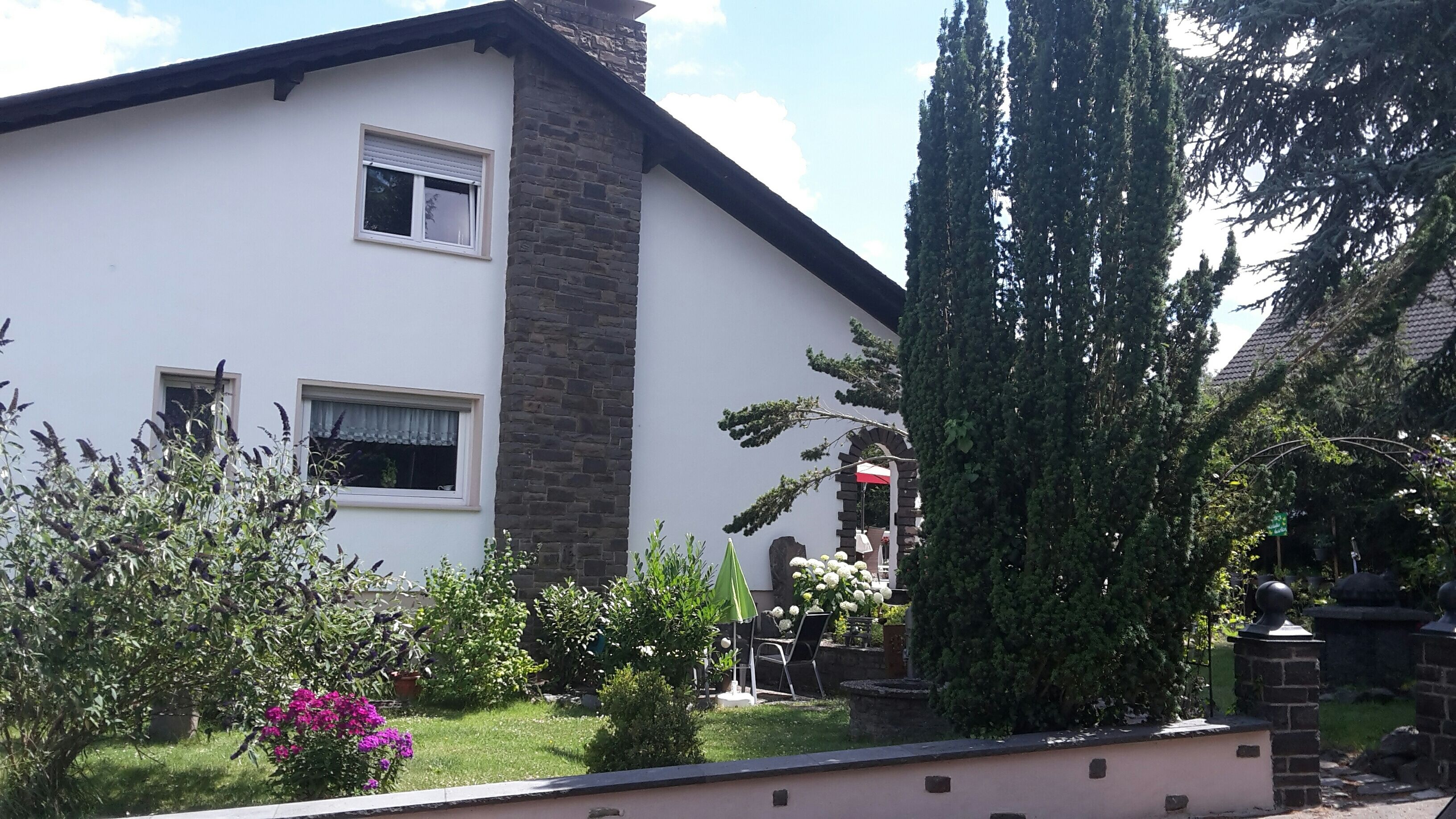 A two-story house with a white facade and a stone chimney. There is a garden with various plants and a tall tree.