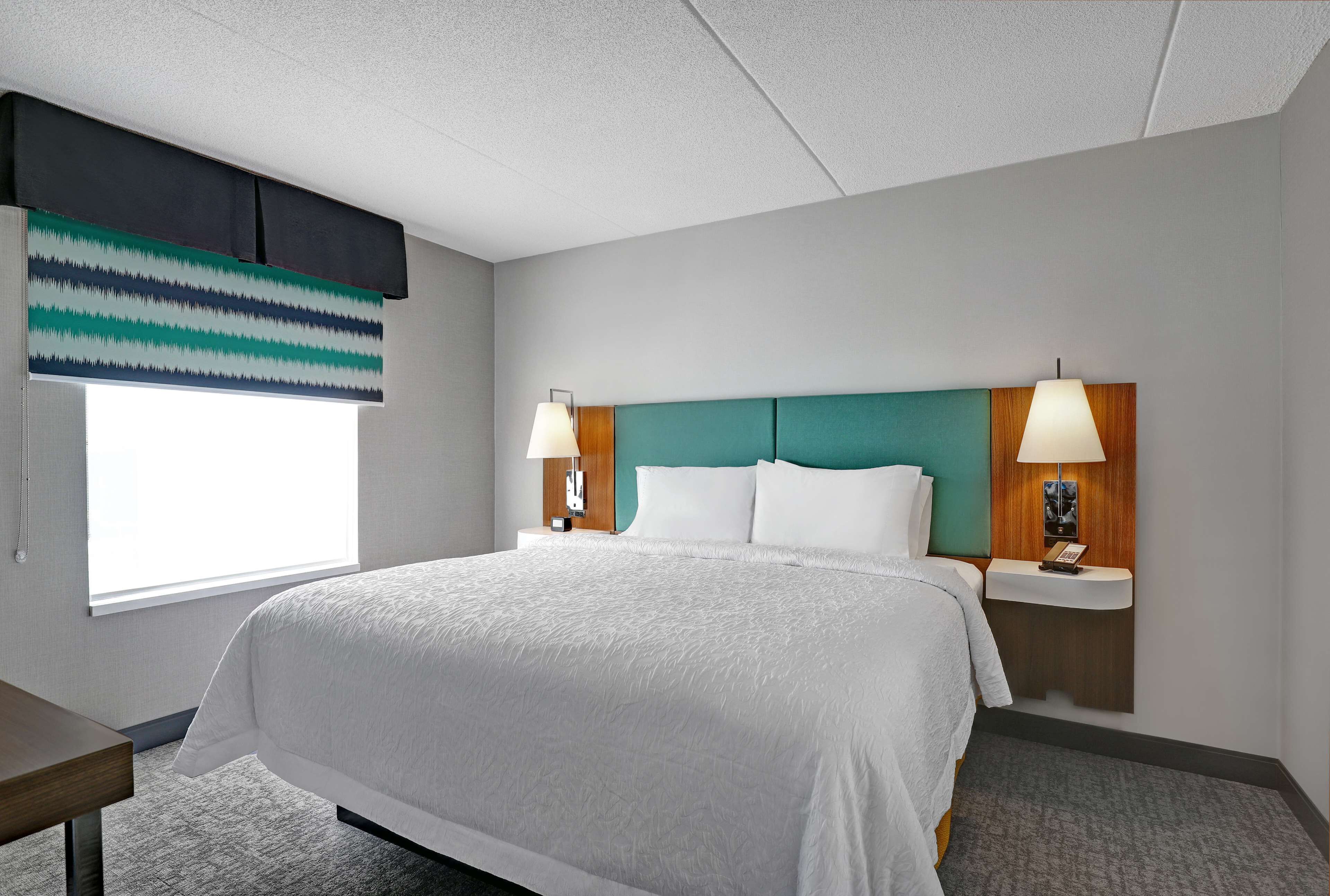 Habitación de hotel con una cama grande, dos mesitas de noche con lámparas, una ventana con persiana a rayas y un cabecero color aguamarina.