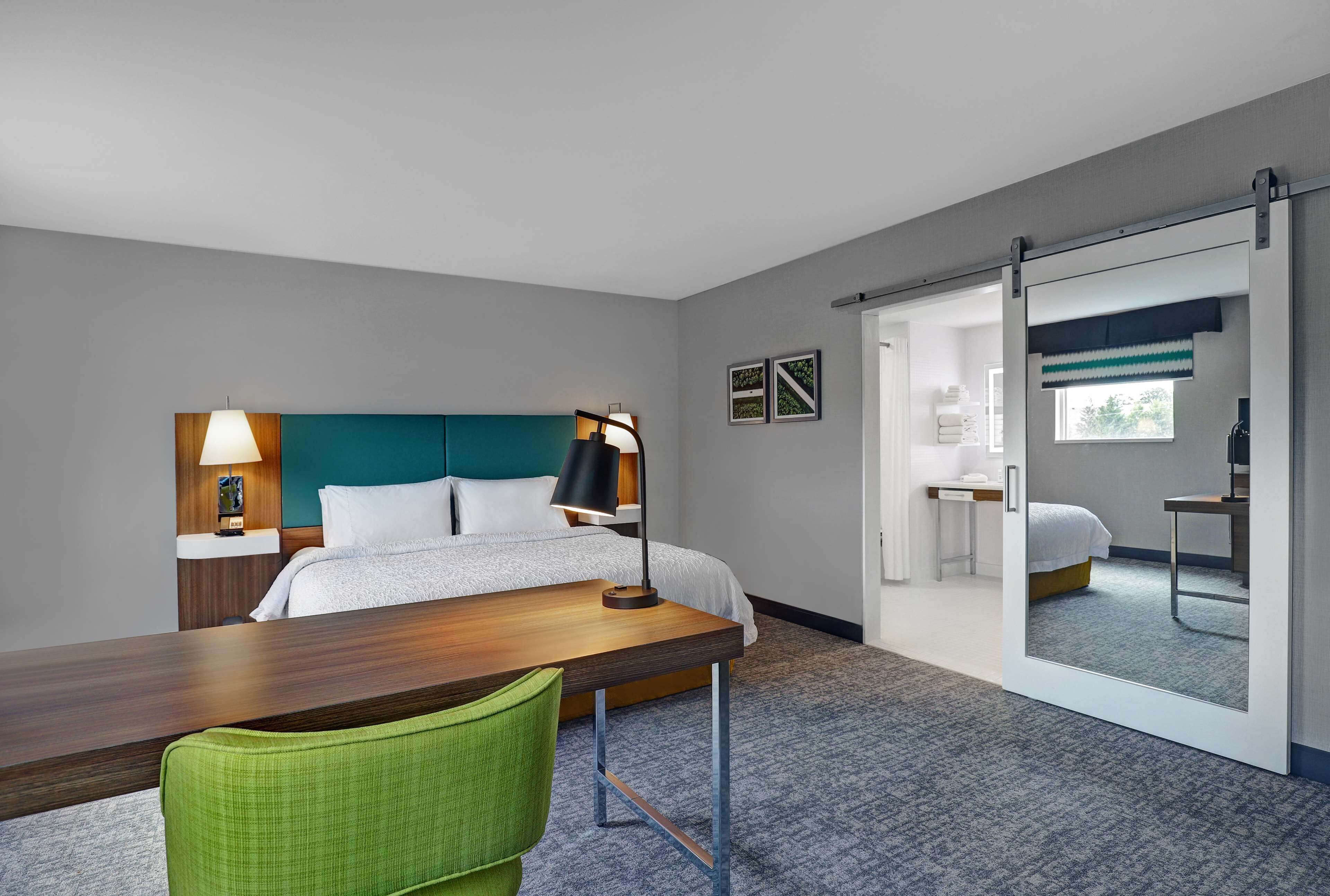 Habitación de hotel con cama, escritorio, silla, lámpara y puerta corredera que da a otra habitación.