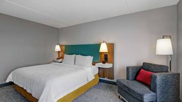 A hotel room with a bed, two bedside lamps, a nightstand, a grey armchair with a red pillow, and a wall-mounted lamp.