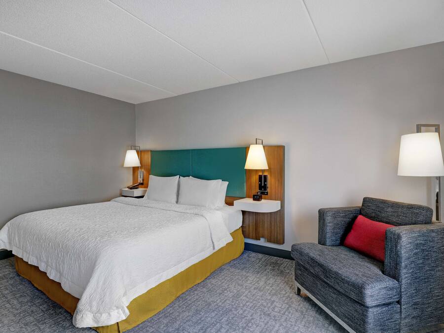 A hotel room with a bed, two bedside lamps, a nightstand, a grey armchair with a red pillow, and a wall-mounted lamp.