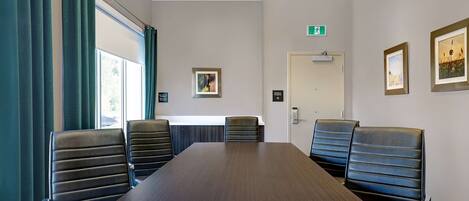 A conference room with a long table and six black leather chairs. There are two windows with green curtains, a door with an exit sign, and framed pictures on the walls.