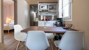 A compact kitchen with a microwave, sink, and modern appliances. There is a dining table with white chairs and a vase of flowers. A bed is visible in the adjacent room. - Olympia's Apartments (Spata-Artemida)