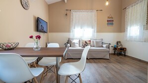 A dining area with a wooden table and white chairs, a sofa with cushions, a clock, and a framed picture on the wall. - Olympia's Apartments (Spata-Artemida)