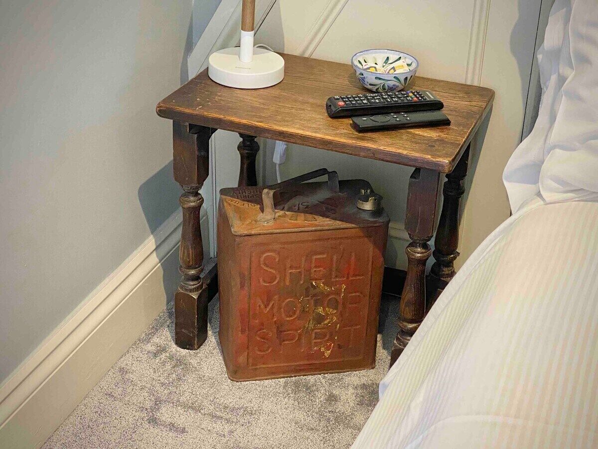 Un comodino in legno con una lampada, un telecomando e una ciotola decorativa. Sotto il tavolo, una tanica vintage per carburante.