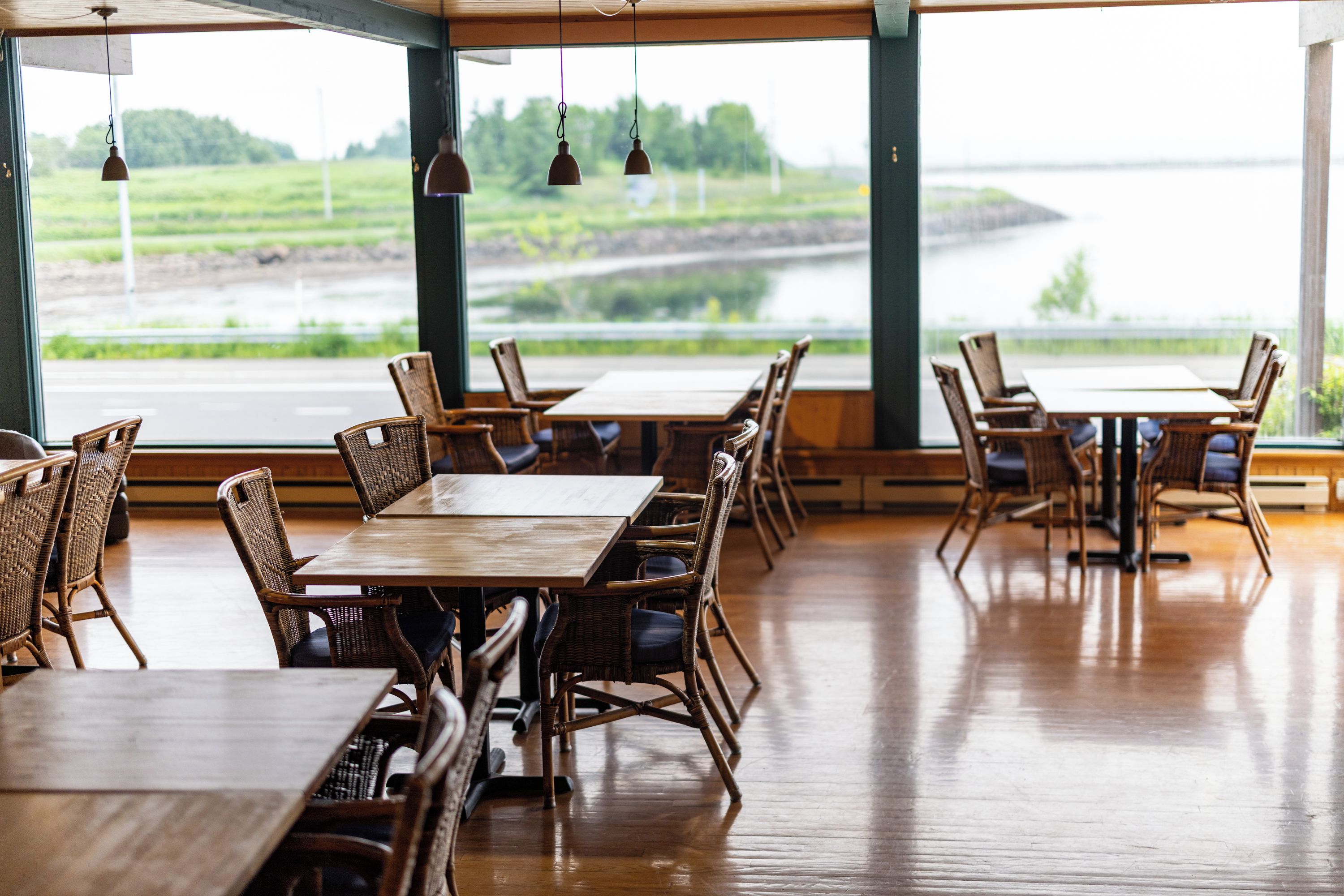 An indoor dining area with wooden tables and wicker chairs, large windows with a view of a river and greenery.