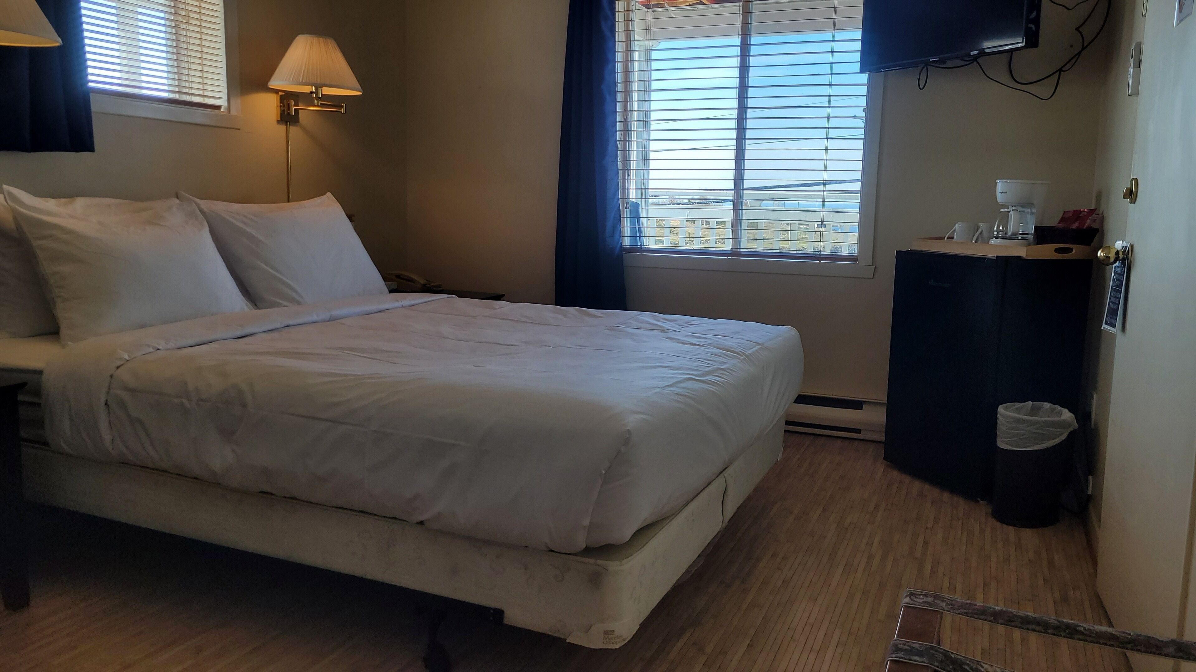 Habitación de hotel con cama, lámparas de noche, ventana con persianas, una mesita con cafetera y un cesto de basura.