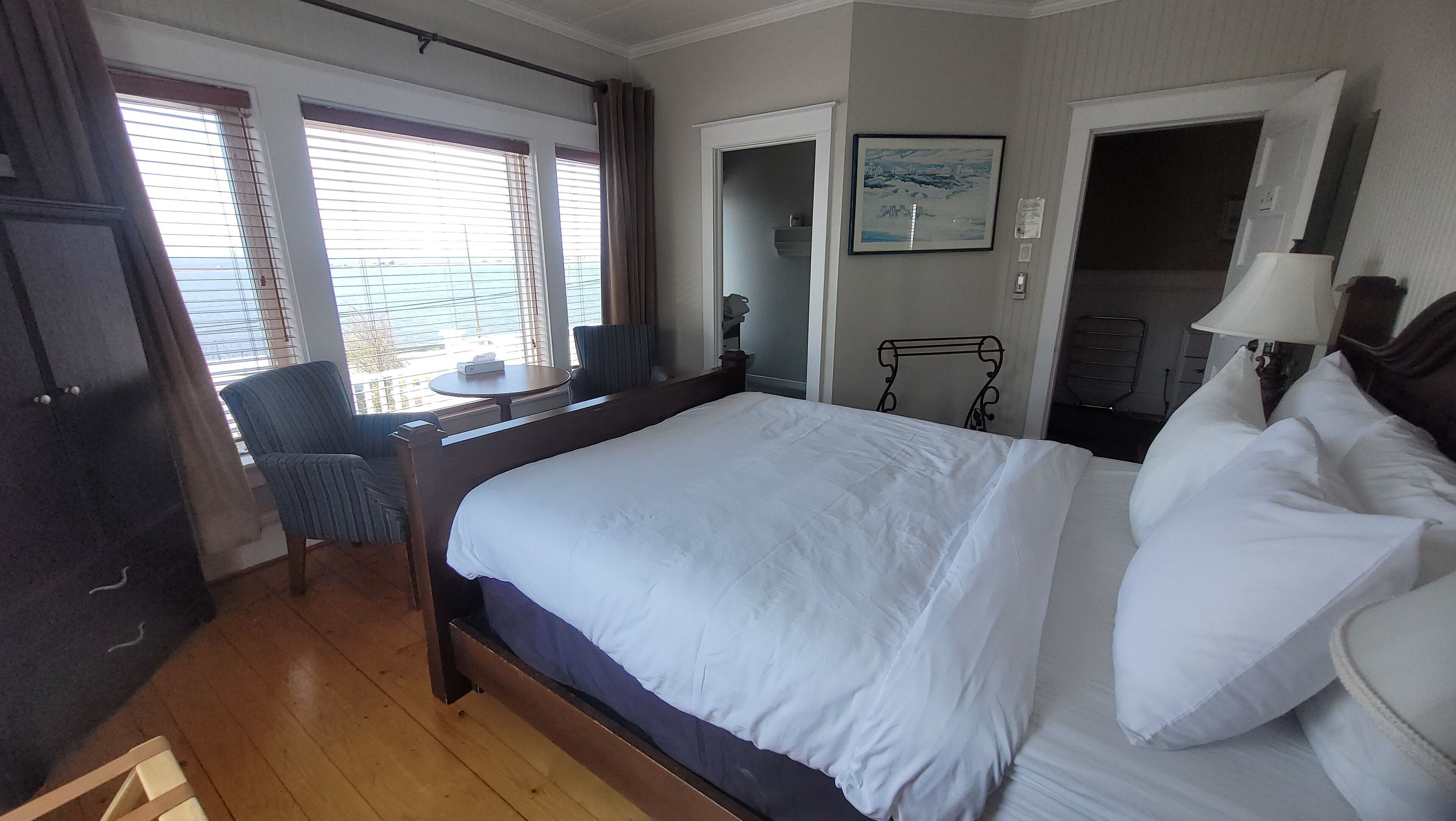 A hotel room with a large bed, a desk with a chair, a window with blinds, a door, and a framed picture on the wall.