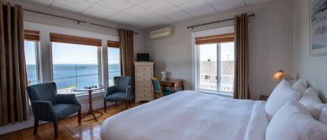 Quarto de hotel com uma cama grande, duas poltronas, uma pequena mesa e uma escrivaninha. Há janelas com cortinas, uma luminária no teto e um ar condicionado fixado na parede.
