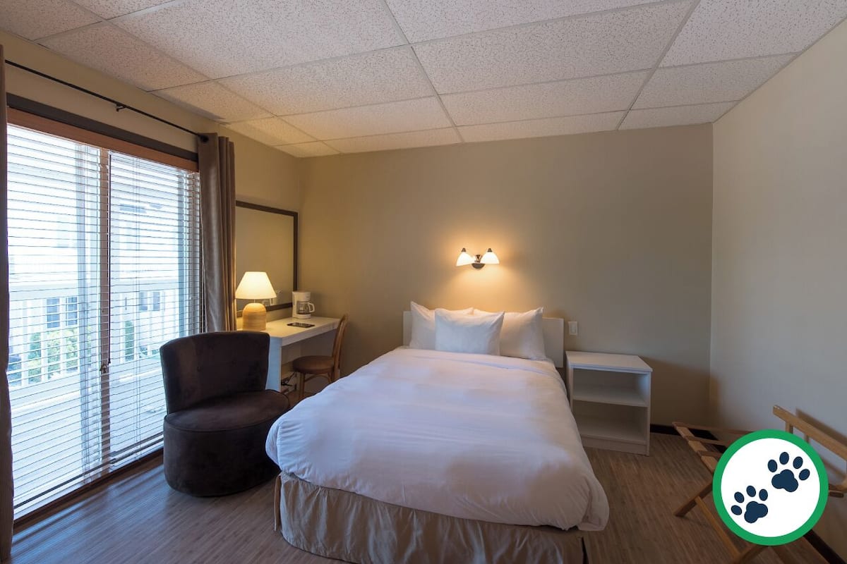 Ein Hotelzimmer mit Bett, Schreibtisch, Stuhl, Lampe und einem Fenster mit Jalousien.