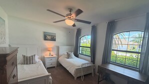 Una camera da letto con due letti, una scrivania, una ventola a soffitto, un comodino con una lampada e una finestra che si affaccia sul verde.
