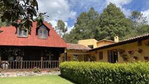 Una casa de dos plantas con techo rojo y fachada amarilla, rodeada de un césped cuidado y setos.