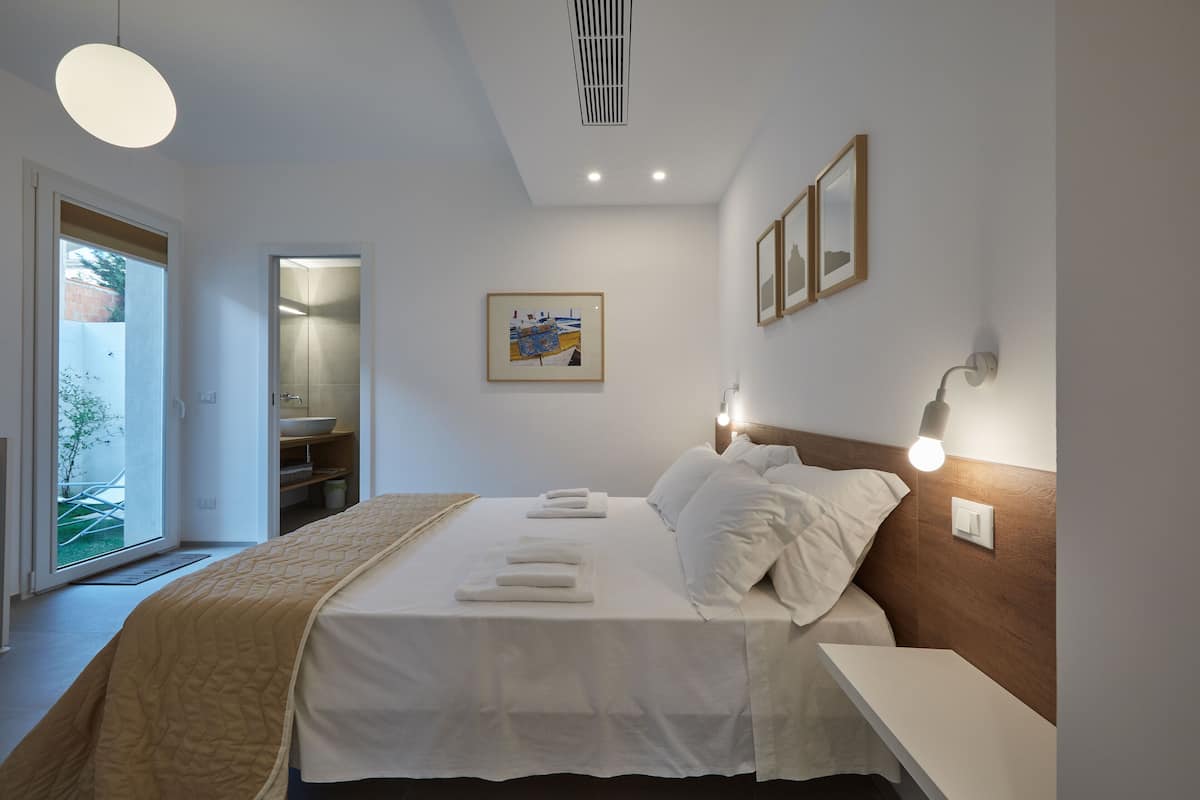 A modern hotel room with a large bed, bedside tables, a headboard with integrated lighting, a framed artwork on the wall, and a view through the door.