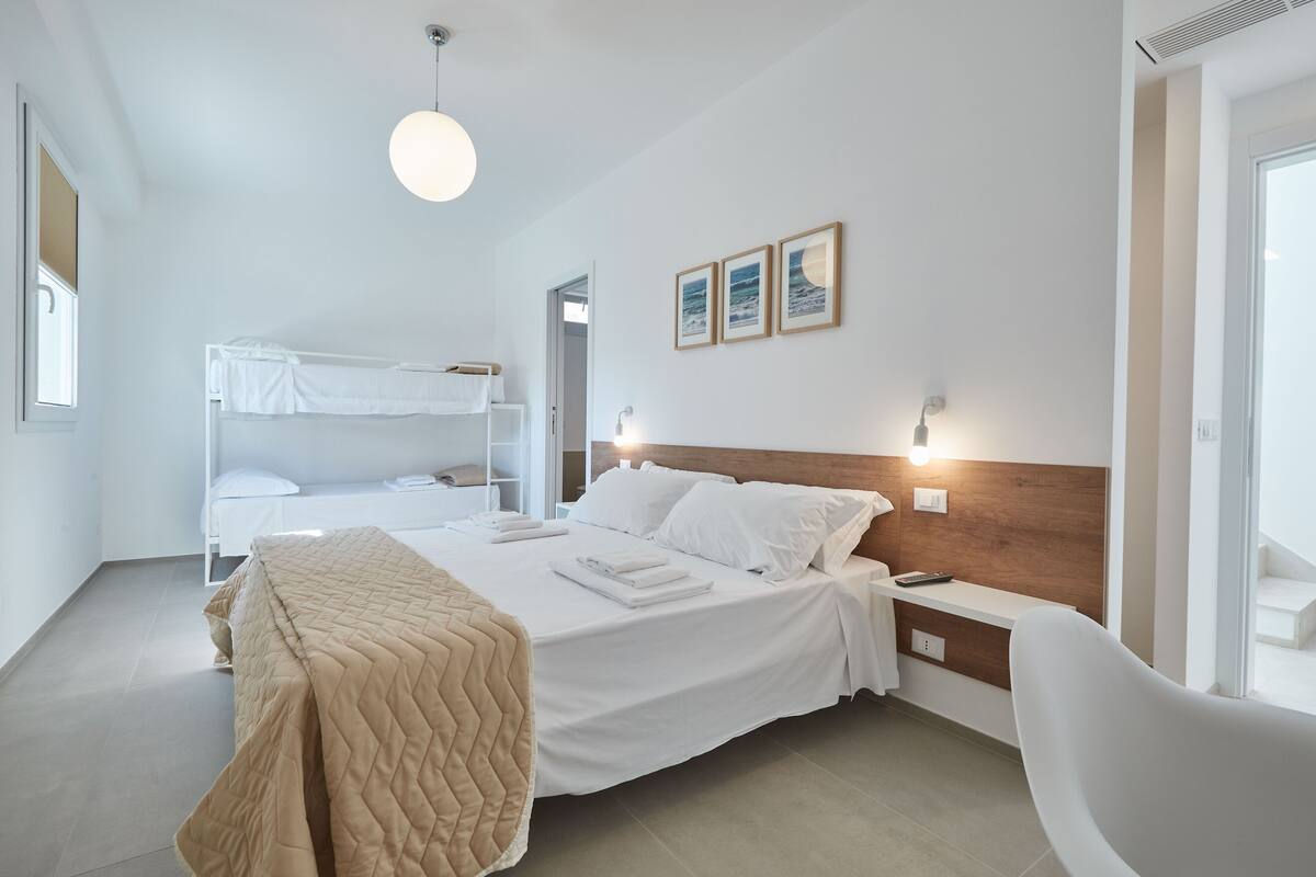 A modern hotel room with a large bed, a smaller bunk bed, a bedside table, and a chair.