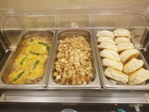 A buffet with three trays of food: a casserole with cheese and green peppers, a tray of diced potatoes, and a tray of biscuits.